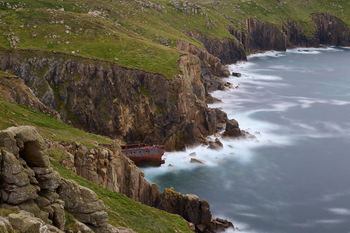 Cornwall shipwreck long exposure This landscape photograph captures a dramatic view of the Cornwall coast, featuring rugged cliffs and the sea in the late afternoon during early summer. The main subject of the image is a shipwreck nestled among the rocks at the base of these cliffs, framed by lush green grass and large boulders. The long exposure technique creates a misty effect over the water, emphasizing the natural beauty of the sea against the striking geological formations of the Cornwall cliffs.